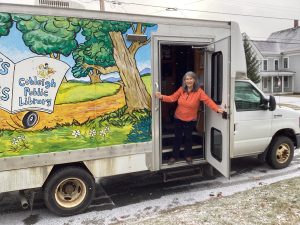 Bookmobile coordinator standing invitingly in doorway of Bookmobile which is painted with a mural of a tree by a road which has a bookmobile travelling down it